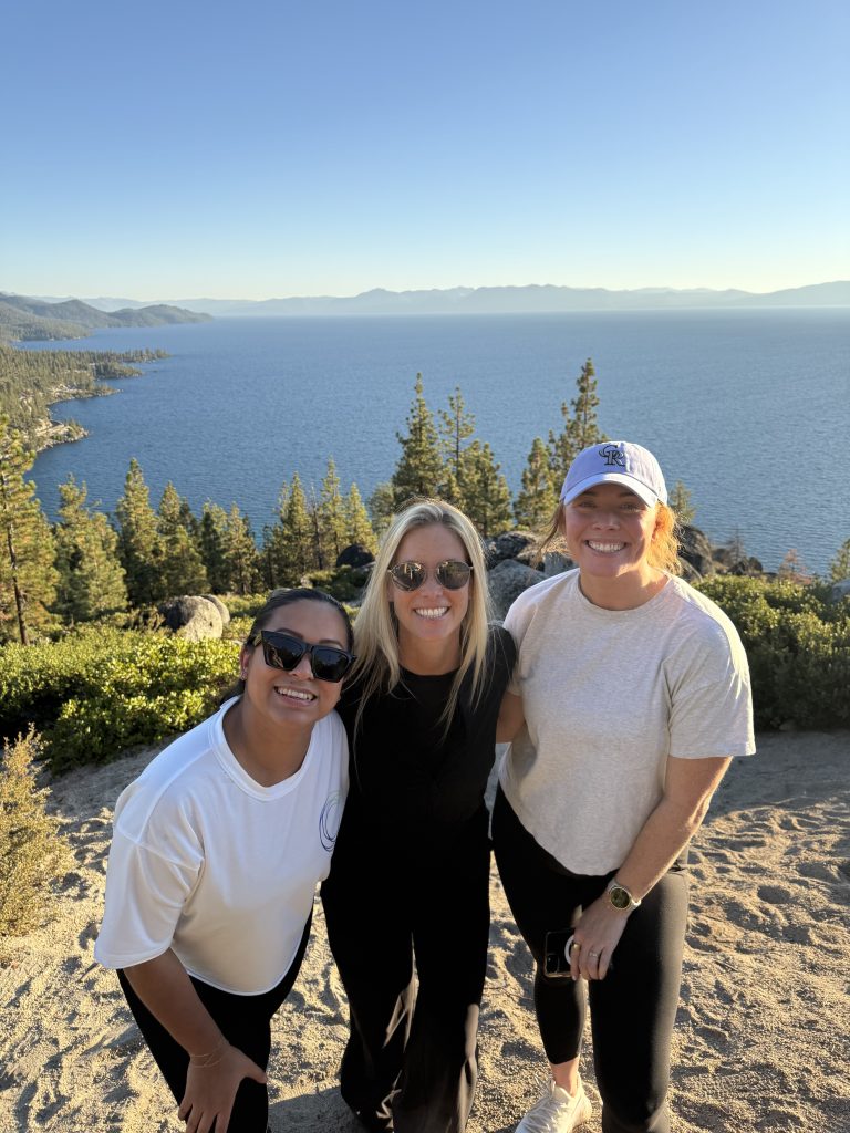 Three women smiling together overlooking a scenic lake and mountains during an outdoor event